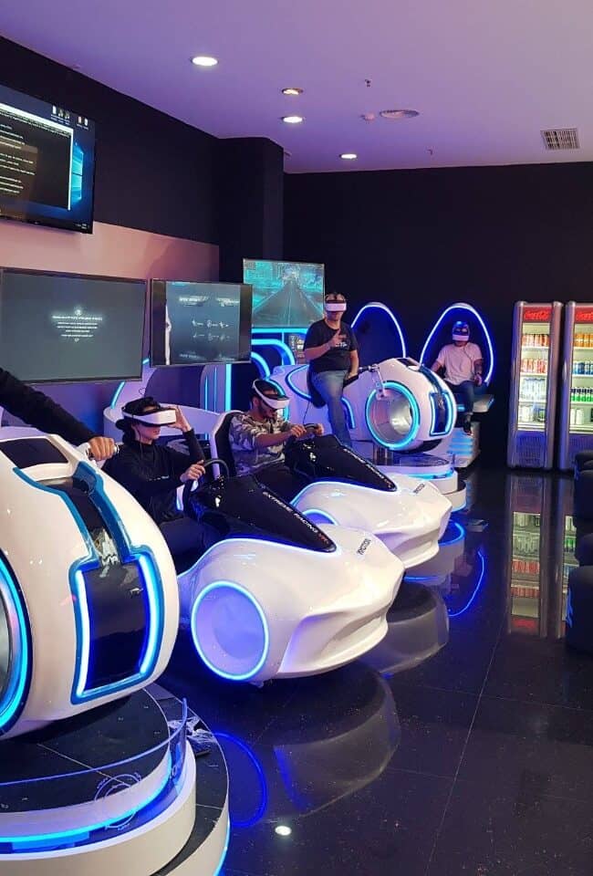 Futuristic virtual reality arcade with players wearing VR headsets and riding simulated racing pods, illuminated by vibrant blue LED lighting, inside an entertainment center.