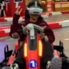 mmexport1736150365286 VR arcade racing game with a young girl wearing a virtual reality headset, sitting in a red go-kart, giving a peace sign, in an indoor entertainment center, colorful background, immersive virtual gaming experience.