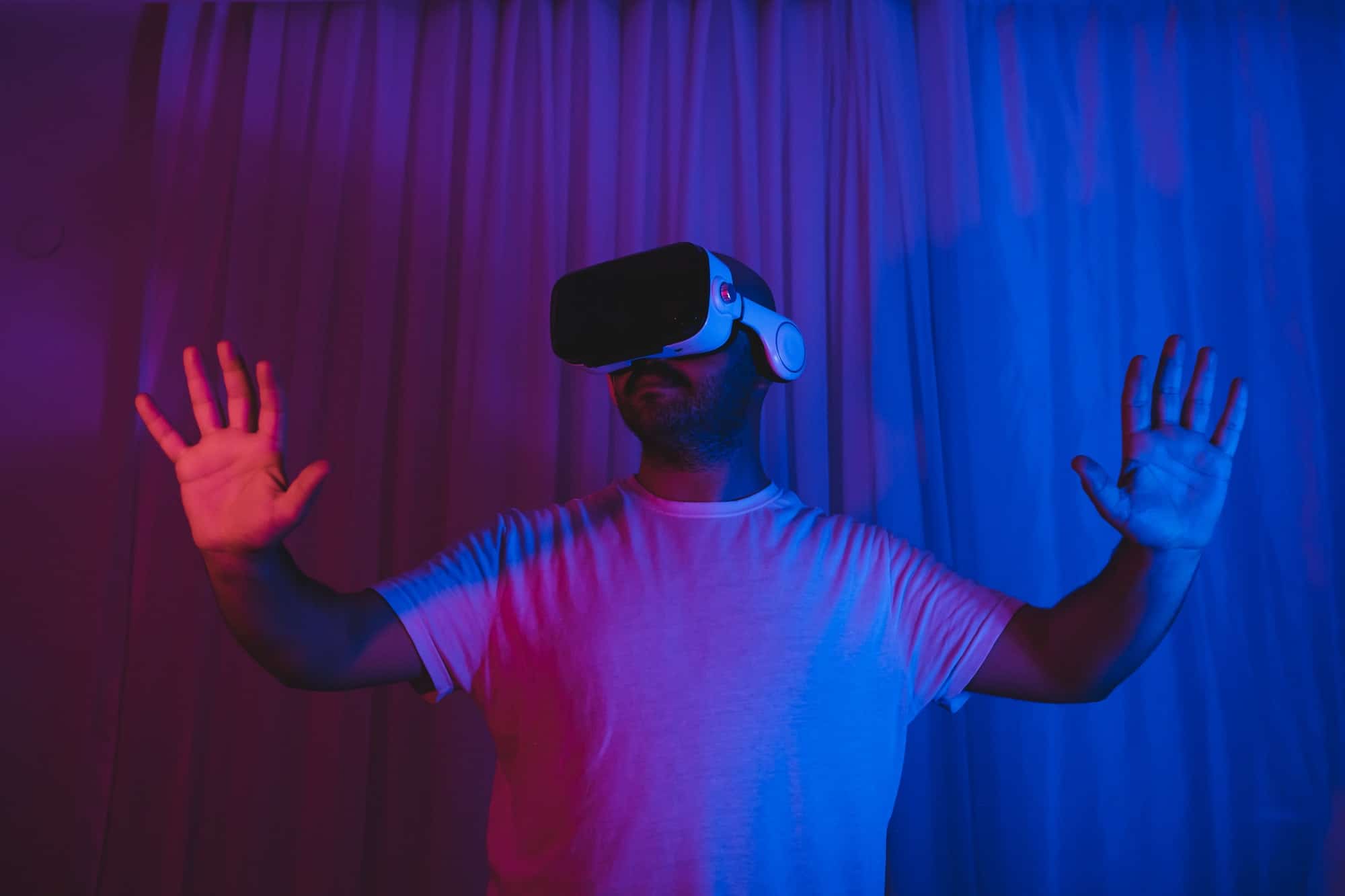 Person using a virtual reality headset with hands raised, immersed in a VR gaming experience, in a dimly lit room with colorful lighting.
