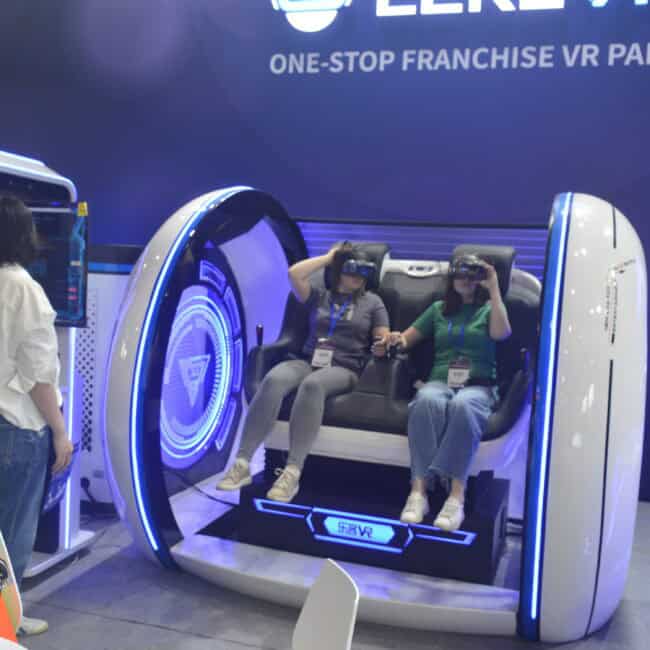 Immersive VR experience at the Lekevrexport booth, showcasing cutting-edge virtual reality technology and entertainment solutions at a trade show. Two women are seated in the VR capsule, wearing headsets, while visitors observe.