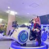 Futuristic arcade game featuring a cosplayer with pink hair and black cat ears sitting on a sci-fi motorcycle arcade machine at a gaming center. VR Motorcycle