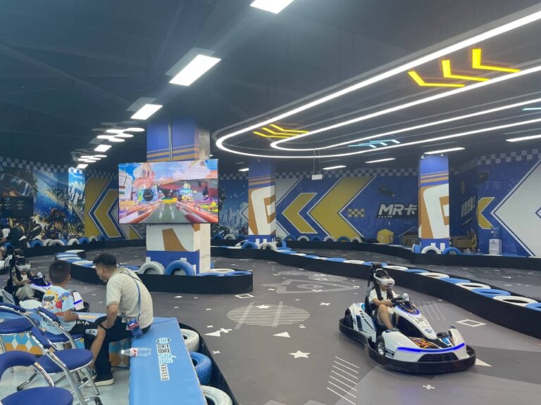 Indoor go-kart racing track with colorful walls and a large screen displaying the track layout, featuring several people and go-karts in action.